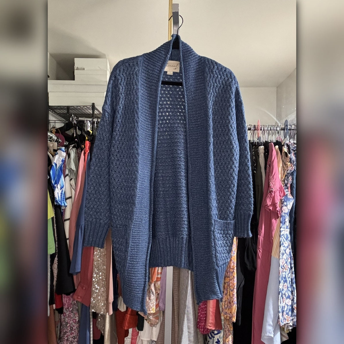 A blue textured knit cardigan sweater hanging on a black hanger in a closet setting with other clothes visible in the background.