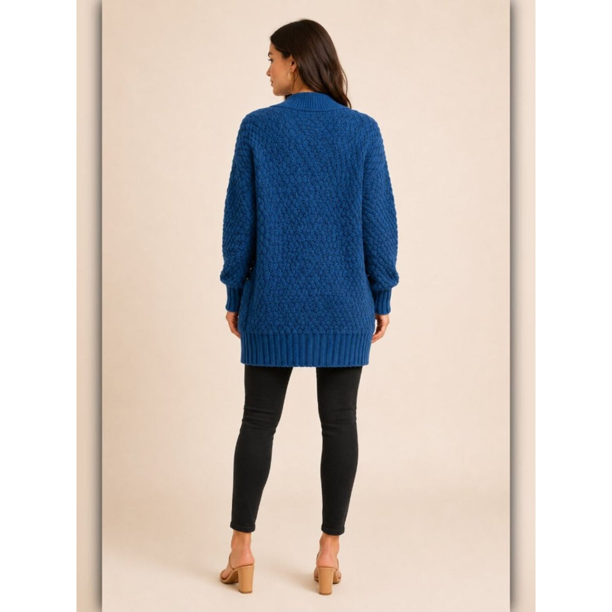 A woman viewed from the back wearing a textured royal blue cable-knit cardigan sweater, black skinny pants, and tan heeled sandals against a solid beige studio background.