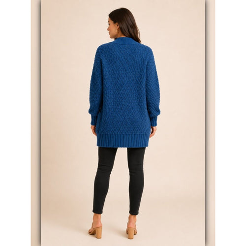 A woman viewed from the back wearing a textured royal blue cable-knit cardigan sweater, black skinny pants, and tan heeled sandals against a solid beige studio background.