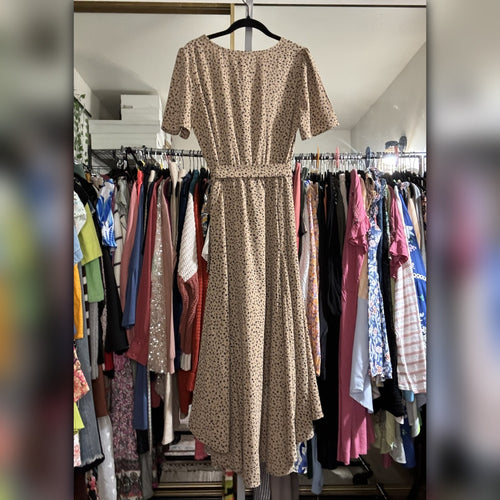 A beige short-sleeve midi dress with a small brown floral print and a matching waist tie, hanging on a black hanger in front of a closet filled with other clothes.