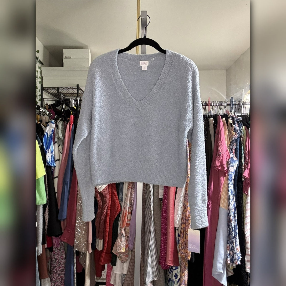 A light gray fuzzy knit long-sleeve V-neck sweater hanging on a black hanger in a closet filled with other clothes.