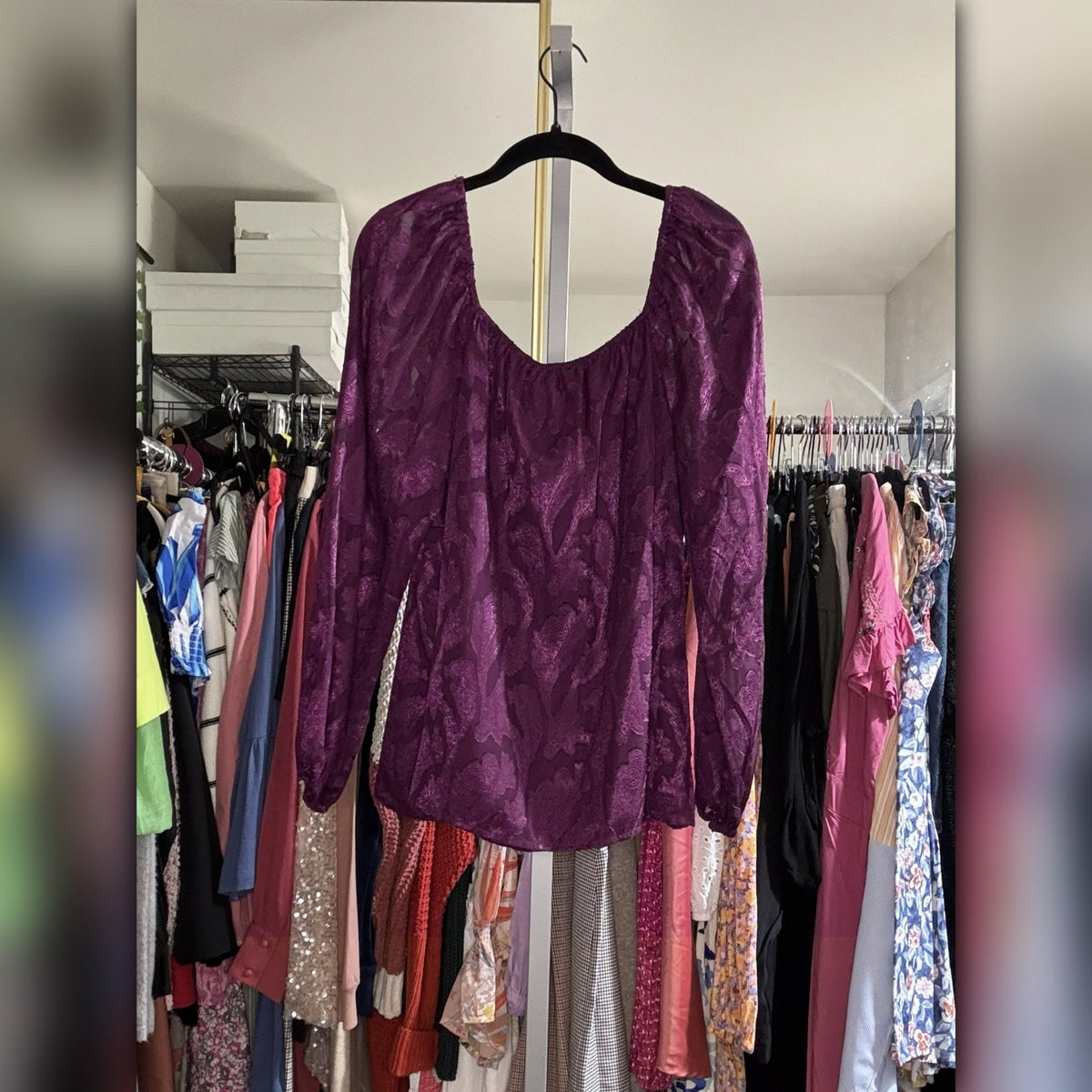 A purple textured long-sleeve top with a scoop neckline hanging on a black hanger in front of a clothing rack.