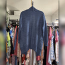 A blue cable-knit sweater with a turtleneck collar and side pockets, hanging on a metal hanger in a closet filled with clothing racks.