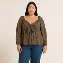 A woman modeling a brown long-sleeve blouse with a blue floral pattern and a front tie detail, paired with blue jeans, against a plain beige background.