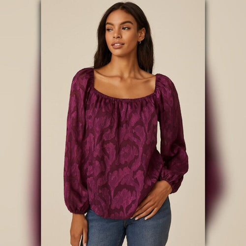 A woman modeling a deep purple long-sleeve blouse with a tonal floral jacquard pattern, a square neckline, and puff sleeves, paired with blue jeans against a beige background.