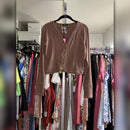 A cropped, long-sleeve cardigan in a dusty mauve-brown color, hanging on a black hanger in a closet filled with other clothes.