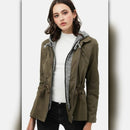 Woman wearing an olive green jacket with a white shirt and black pants on a light gray background