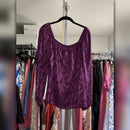 A purple velvet long-sleeve top with a wavy jacquard pattern, hanging on a black hanger in a closet with clothing racks visible in the background.