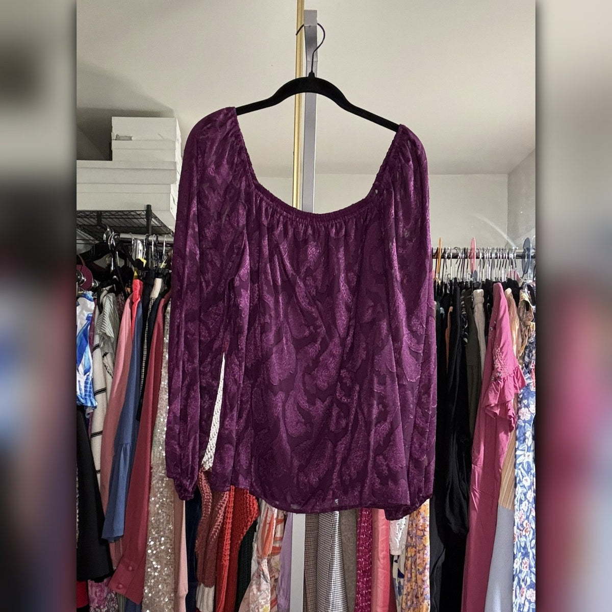 A purple velvet long-sleeve top with a wavy jacquard pattern, hanging on a black hanger in a closet with clothing racks visible in the background.