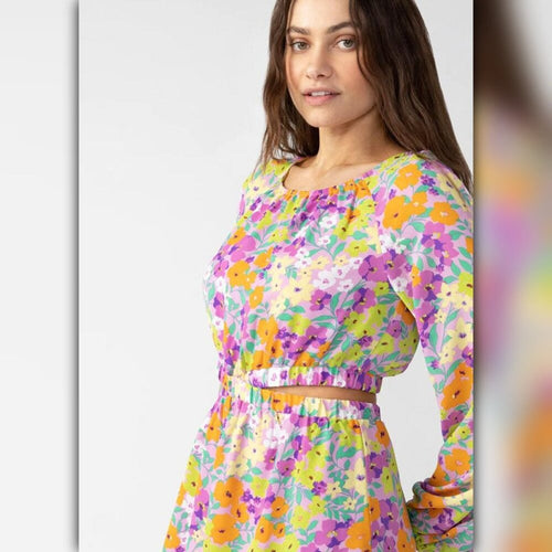 A woman wearing a long-sleeve floral dress with a yellow, purple, orange, and green print, featuring a gathered waist and a ruffled neckline, set against a light gray studio background.
