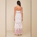 A woman viewed from the back wearing a white spaghetti-strap maxi dress with a pink and green floral print and a ruffled hem, set against a solid beige studio background.