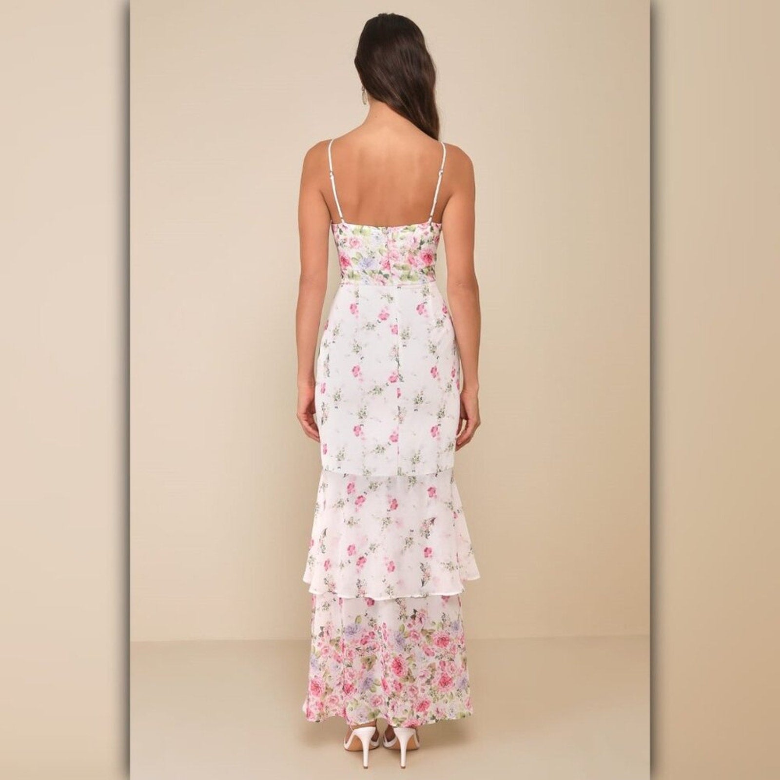 A woman viewed from the back wearing a white spaghetti-strap maxi dress with a pink and green floral print and a ruffled hem, set against a solid beige studio background.