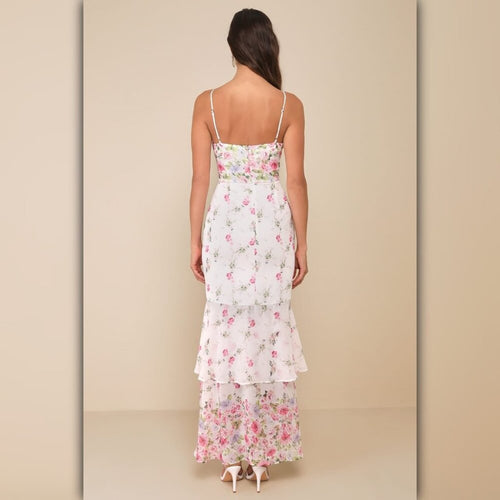 A woman viewed from the back wearing a white spaghetti-strap maxi dress with a pink and green floral print and a ruffled hem, set against a solid beige studio background.