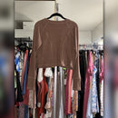 A cropped, long-sleeve ribbed knit top in a warm taupe-brown color, hanging on a black hanger in front of a clothing rack filled with various garments.