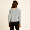 A woman viewed from the back wearing a light gray fuzzy long-sleeve sweater and black pants, standing against a plain off-white background.