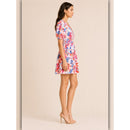 A woman modeling a short puff-sleeve mini dress with a vibrant red, blue, and pink floral print on a white base, paired with beige high-heeled sandals, set against a solid beige studio background.