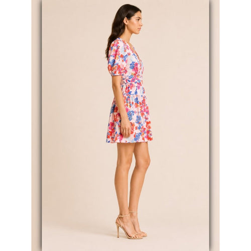 A woman modeling a short puff-sleeve mini dress with a vibrant red, blue, and pink floral print on a white base, paired with beige high-heeled sandals, set against a solid beige studio background.