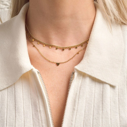 Close-up of a person wearing a gold necklace with a heart pendant on a light-colored shirt.