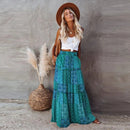 Woman in a white top and green patterned skirt standing next to pampas grass against a textured wall.