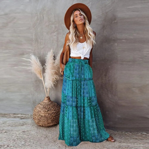 Woman in a white top and green patterned skirt standing next to pampas grass against a textured wall.
