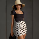 Woman wearing a black top and white skirt with black polka dots, holding a black handbag against a gray background.