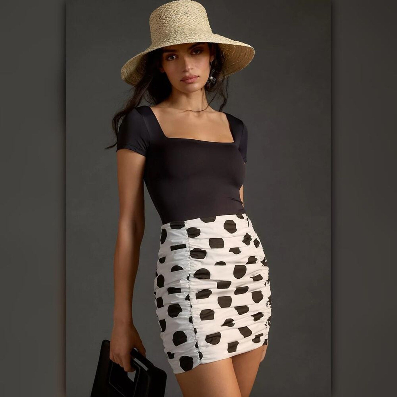 Woman wearing a black top and white skirt with black polka dots, holding a black handbag against a gray background.