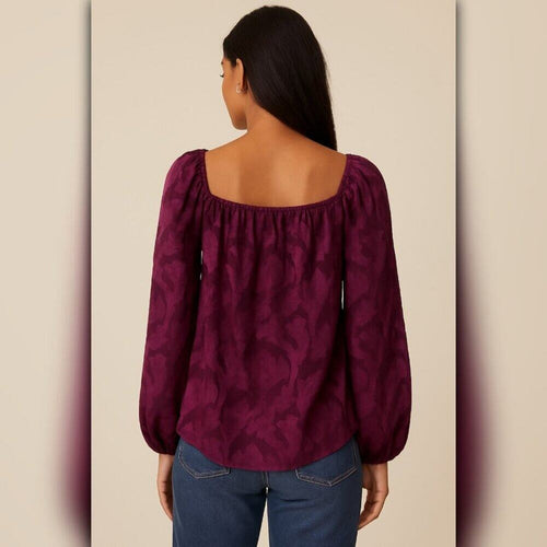 A woman shown from the back wearing a deep burgundy off-the-shoulder long-sleeve blouse with a subtle tonal damask pattern, paired with blue jeans, against a neutral beige background.