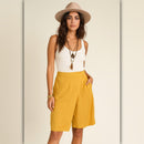 A woman modeling a pair of mustard yellow high-waisted shorts with a white ribbed tank top, layered necklaces, and a beige wide-brim hat, set against a warm beige studio background.