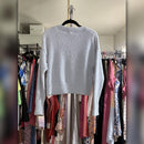A light gray textured knit long-sleeve crop top hanging on a black hanger in front of a closet rack filled with colorful clothing.