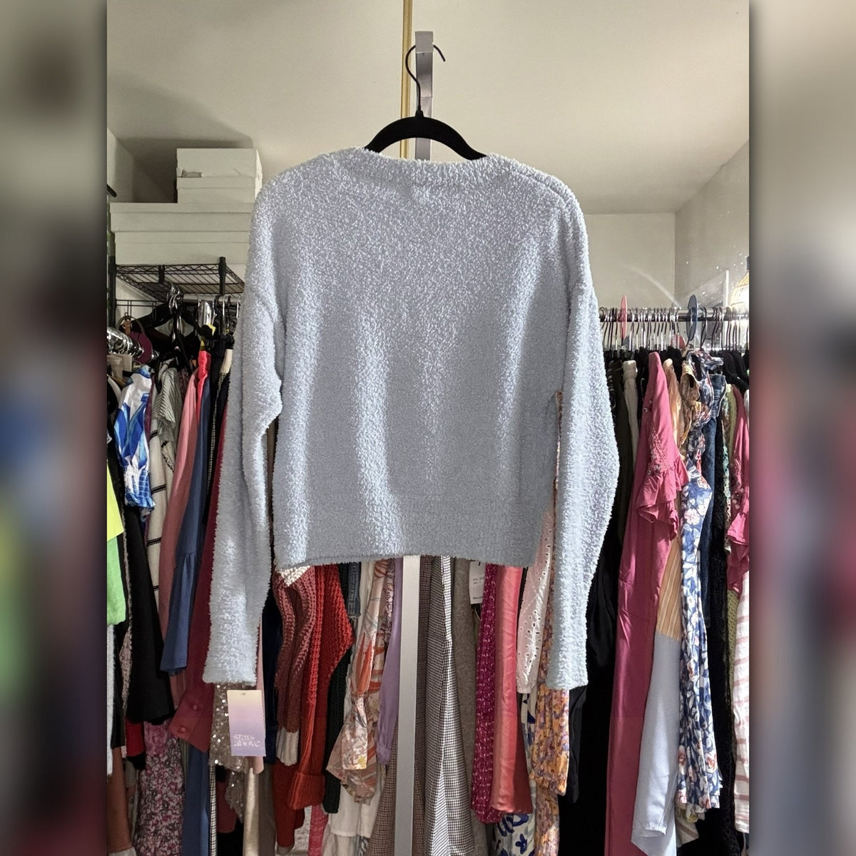 A light gray textured knit long-sleeve crop top hanging on a black hanger in front of a closet rack filled with colorful clothing.