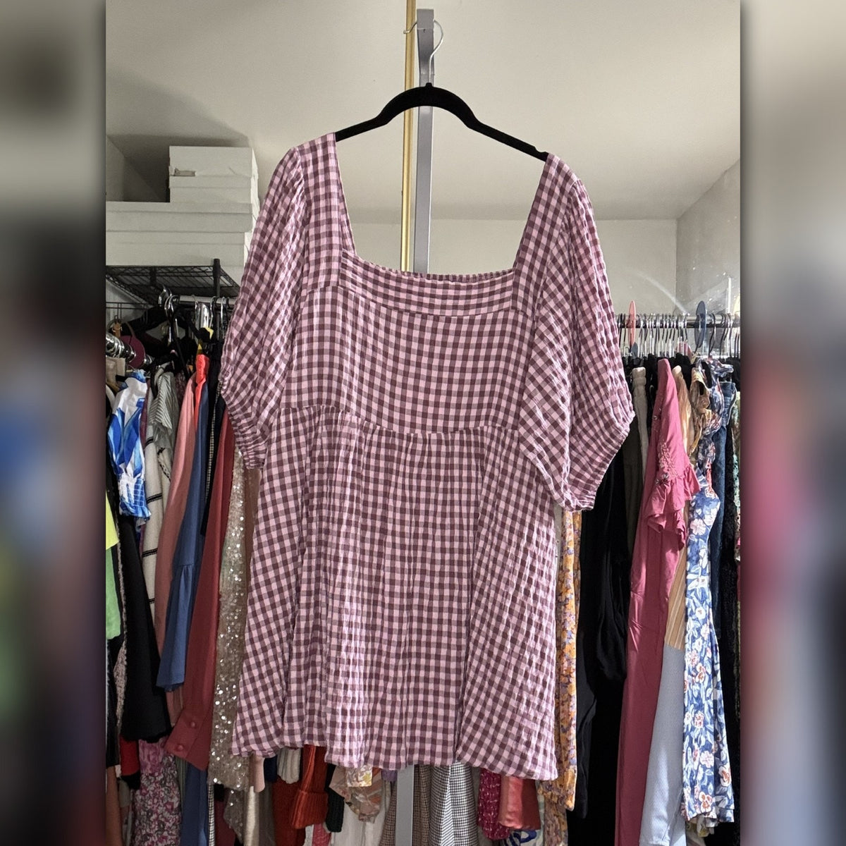 A pink and white gingham short-sleeve top hanging on a black hanger in a closet area with other clothes visible in the background.