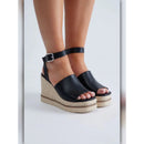 A pair of women's black leather wedge sandals with espadrille-style woven platform soles and ankle straps, shown on a model's feet against a light gray studio background.