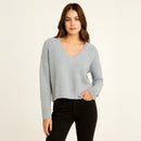 A woman modeling a light gray fuzzy long-sleeve V-neck sweater with black pants, standing against a plain off-white studio background.