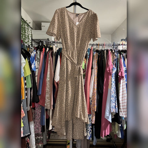 A beige short-sleeve wrap-style dress with a small black floral print hangs on a black hanger in a clothing store, surrounded by racks of various colorful garments.