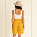 A woman shown from the back wearing a beige wide-brim hat, a white ribbed tank top, and mustard yellow shorts against a light cream background.
