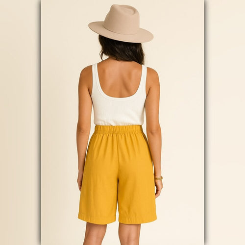 A woman shown from the back wearing a beige wide-brim hat, a white ribbed tank top, and mustard yellow shorts against a light cream background.