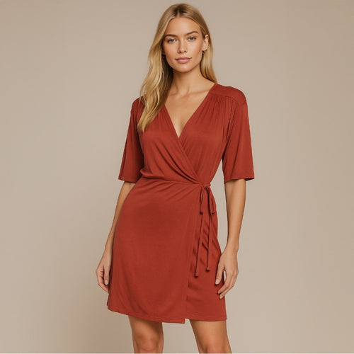 A woman modeling a rust-colored wrap mini dress with short sleeves and a tie waist, set against a solid beige studio background.