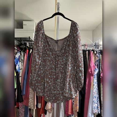 A brown long-sleeve blouse with a light blue floral pattern, hanging on a black hanger in front of a closet filled with other clothes.