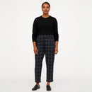A woman stands against a plain light gray studio background wearing a black long-sleeve top and dark navy plaid cropped trousers with white windowpane lines, paired with black loafers.