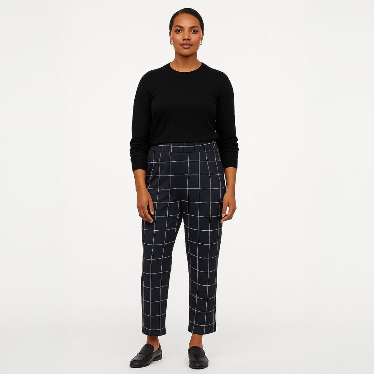 A woman stands against a plain light gray studio background wearing a black long-sleeve top and dark navy plaid cropped trousers with white windowpane lines, paired with black loafers.