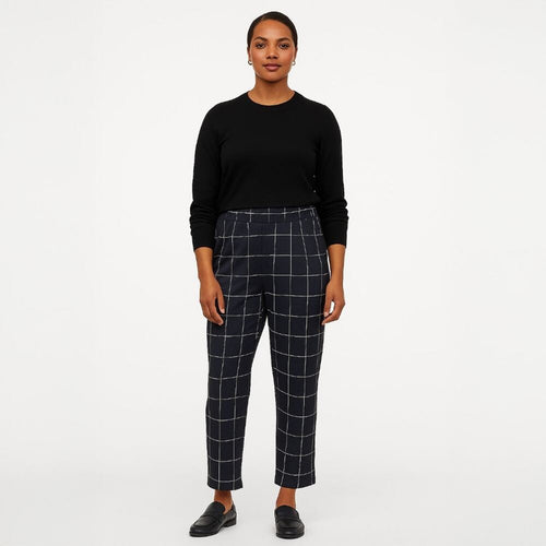 A woman stands against a plain light gray studio background wearing a black long-sleeve top and dark navy plaid cropped trousers with white windowpane lines, paired with black loafers.