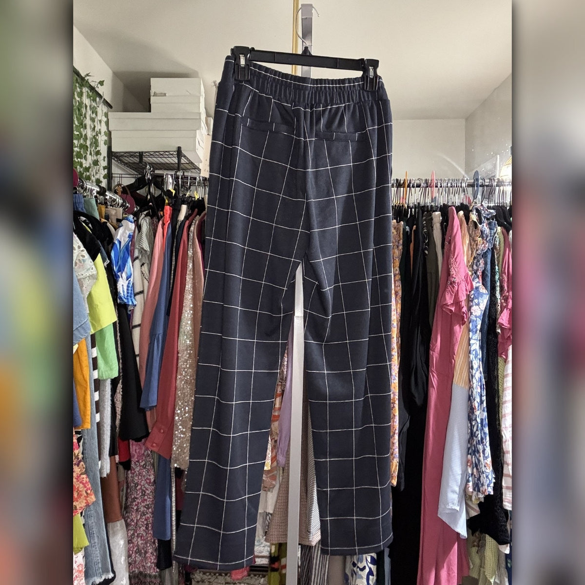 A pair of dark gray wide-leg trousers with a white windowpane grid pattern, hanging on a black hanger in a closet filled with clothing racks.
