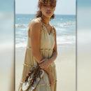 Woman in a beige dress holding sandals on a beach with ocean in the background