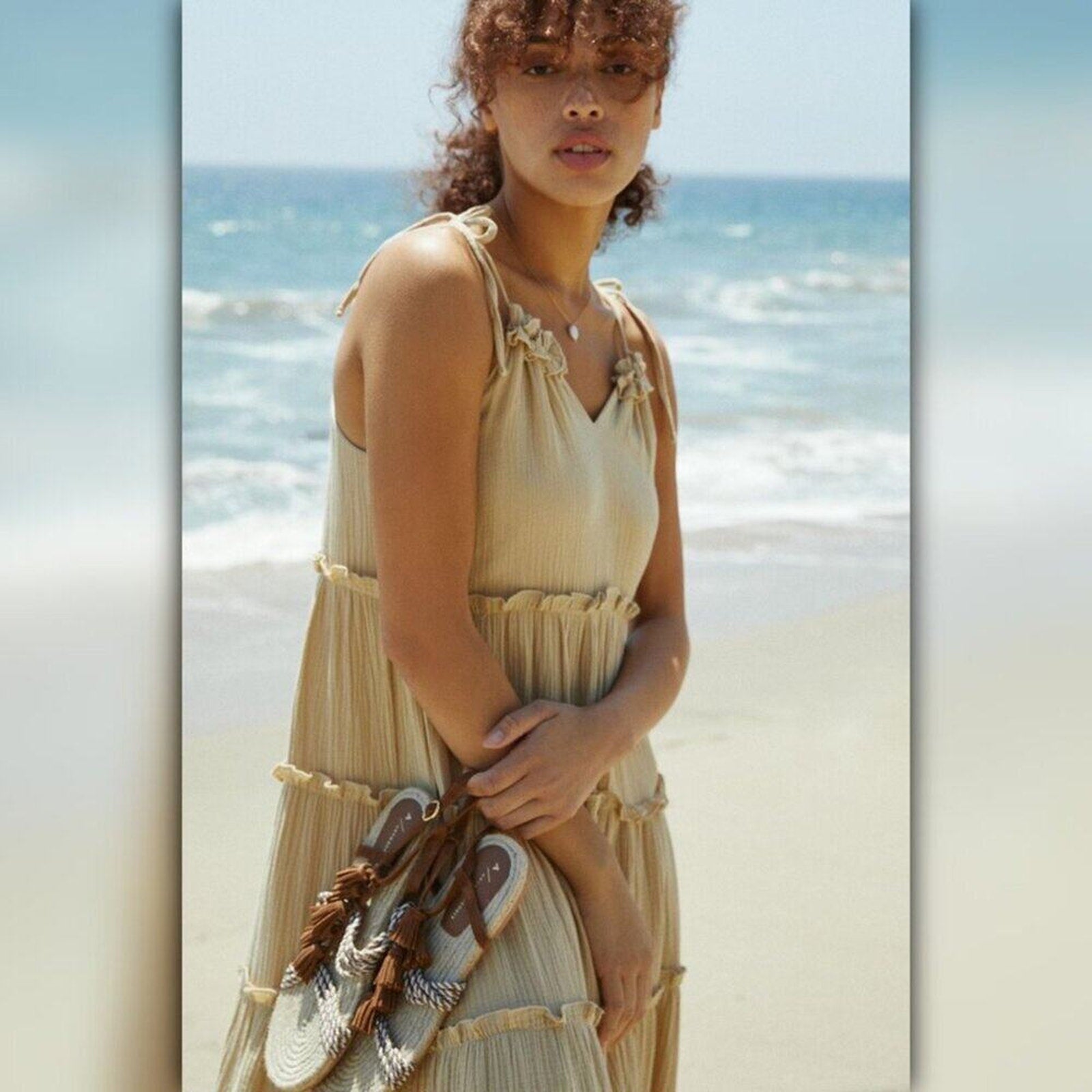 Woman in a beige dress holding sandals on a beach with ocean in the background