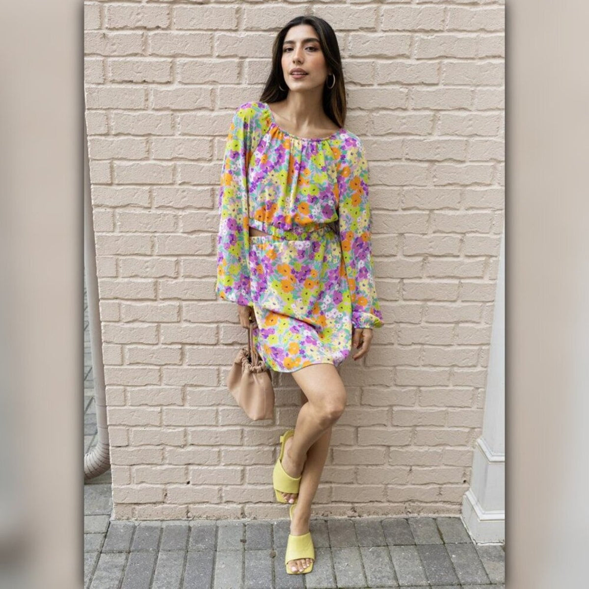 A woman stands against a light brick wall wearing a colorful floral long-sleeve mini dress with a matching belt, holding a small beige handbag and wearing yellow slide sandals.