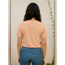 Person wearing a peach t-shirt and blue jeans against a neutral wall with a plant.