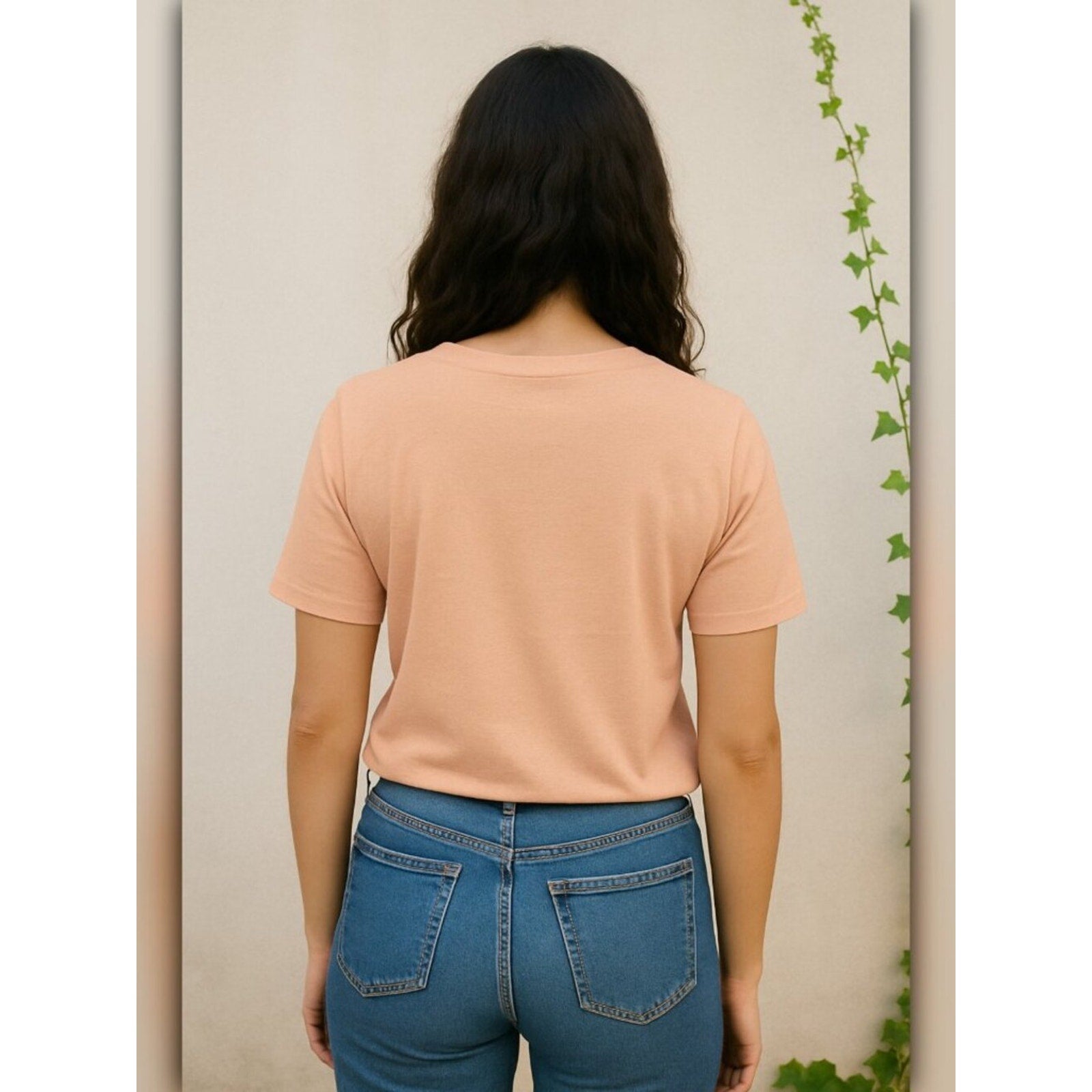 Person wearing a peach t-shirt and blue jeans against a neutral wall with a plant.