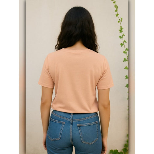 Person wearing a peach t-shirt and blue jeans against a neutral wall with a plant.