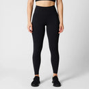 A model wearing ribbed black high-waisted leggings, a black sports bra, and black sneakers, standing against a light gray studio background.