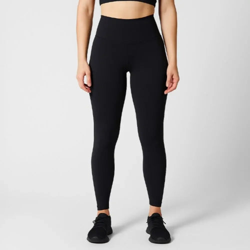 A model wearing ribbed black high-waisted leggings, a black sports bra, and black sneakers, standing against a light gray studio background.